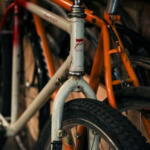Close-up of a white vintage bicycle head tube and front fork with a red logo, with an orange bike in the background in dim storage.