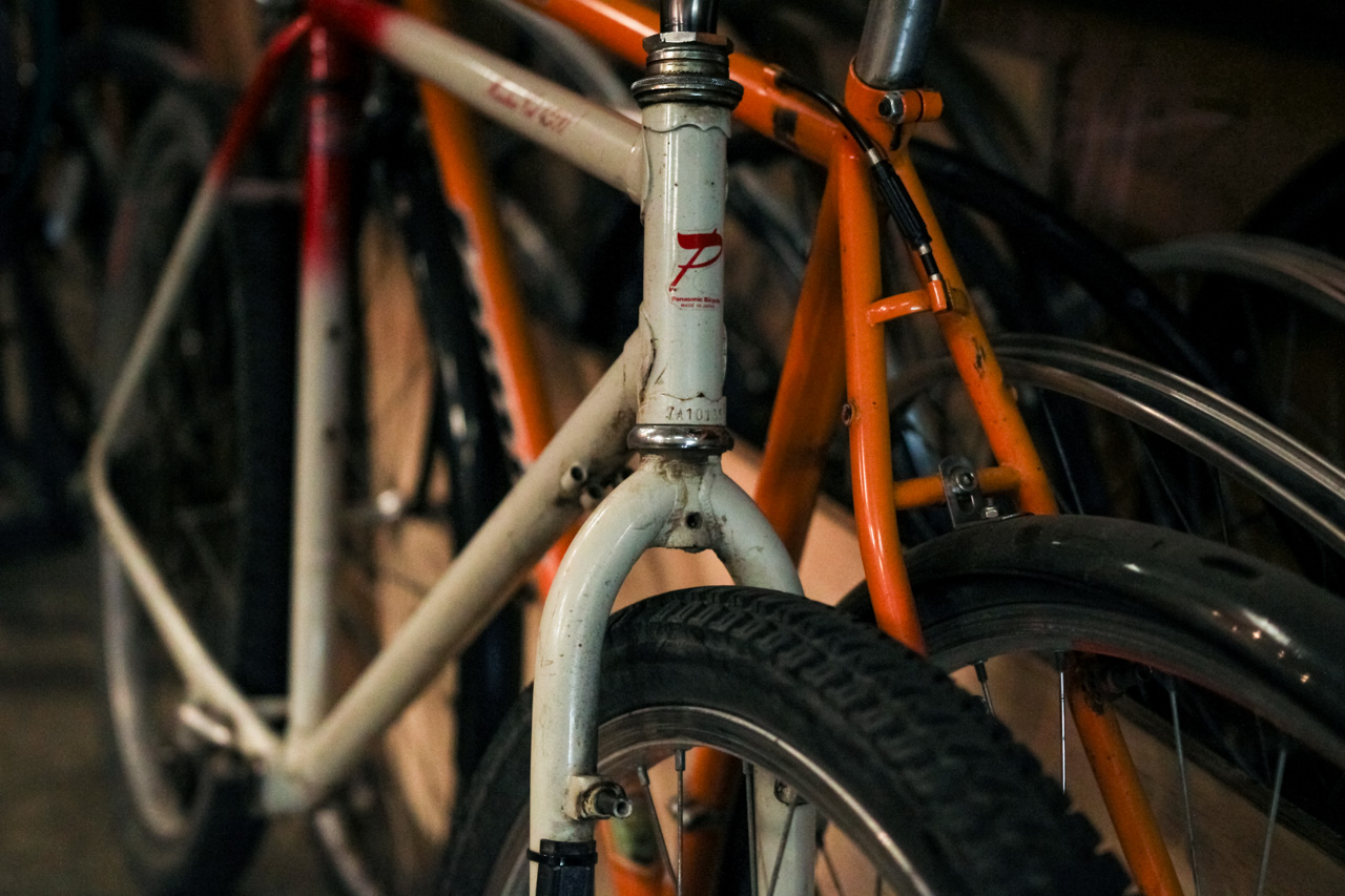 Close-up of a white vintage bicycle head tube and front fork with a red logo, with an orange bike in the background in dim storage.