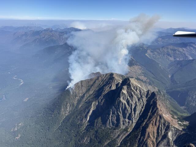 Aerial view of rugged mountains with a tall plume of white smoke rising from a volcano, seen from an airplane window.
