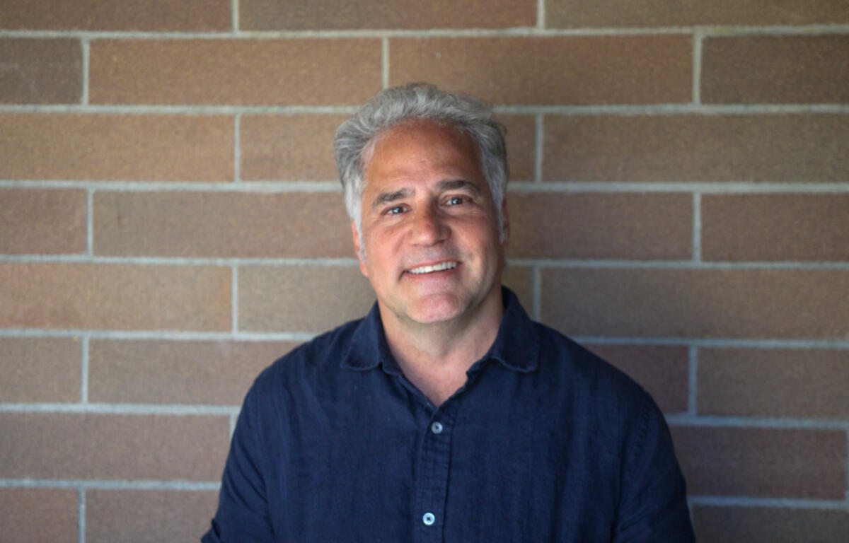 Smiling man with gray hair wearing a dark blue shirt, standing against a brick wall.