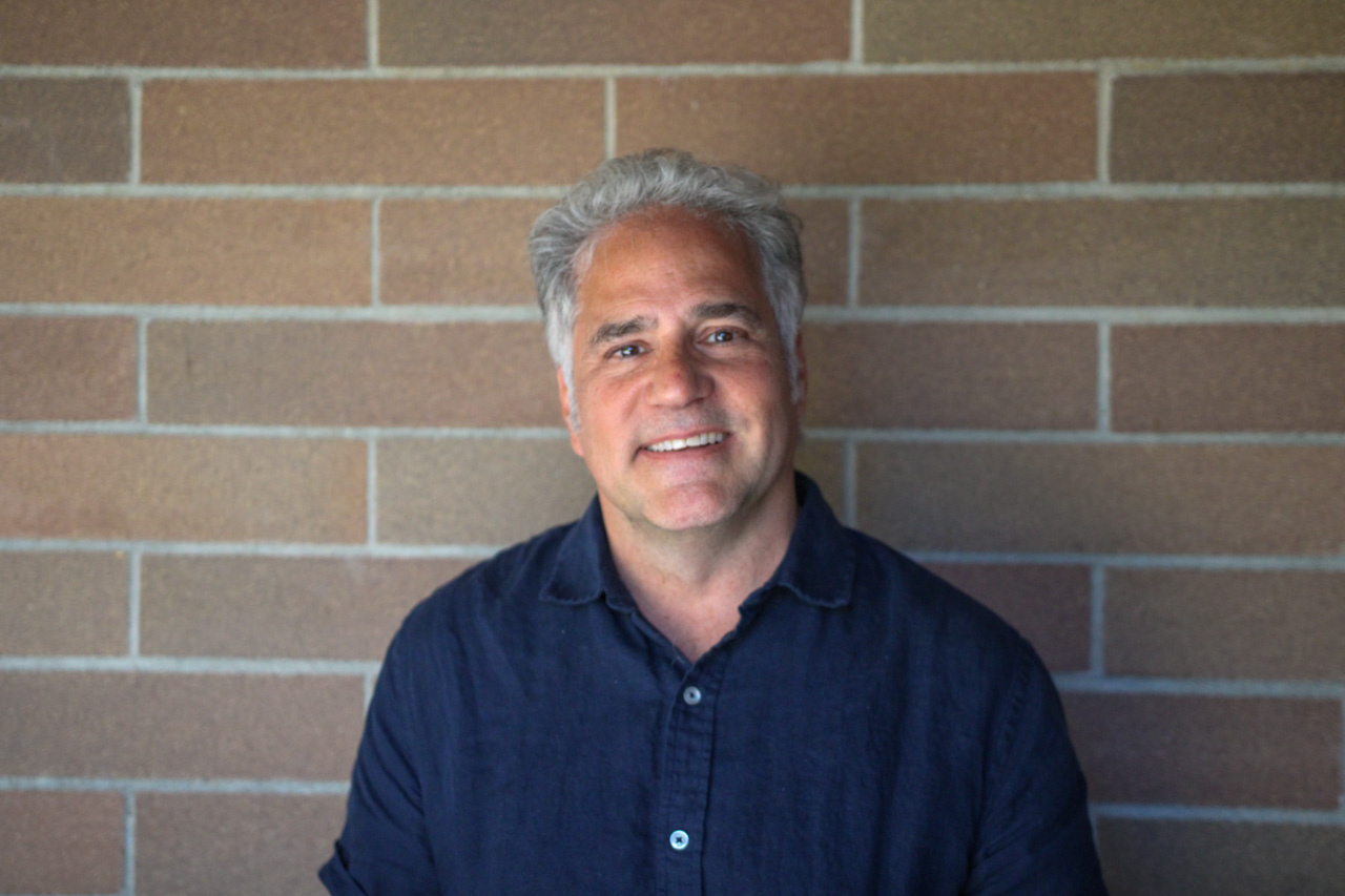 Smiling man with gray hair wearing a dark blue shirt, standing against a brick wall.