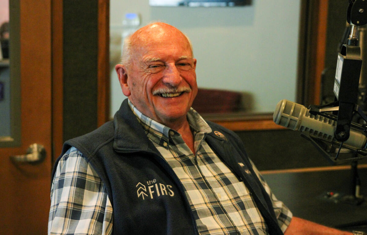 Smiling older man in a plaid shirt and navy vest sits in a radio studio, speaking into a large microphone.