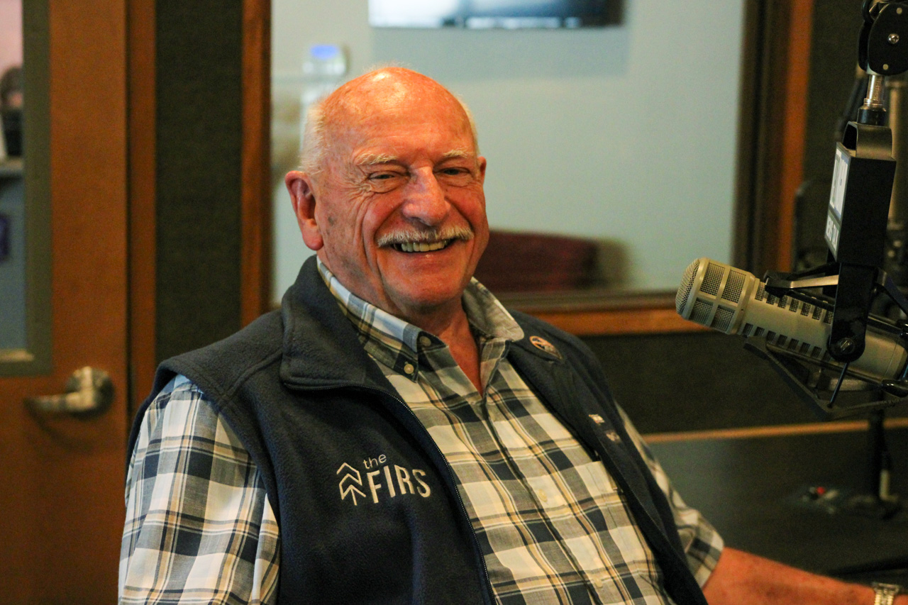 Smiling older man in a plaid shirt and navy vest sits in a radio studio, speaking into a large microphone.