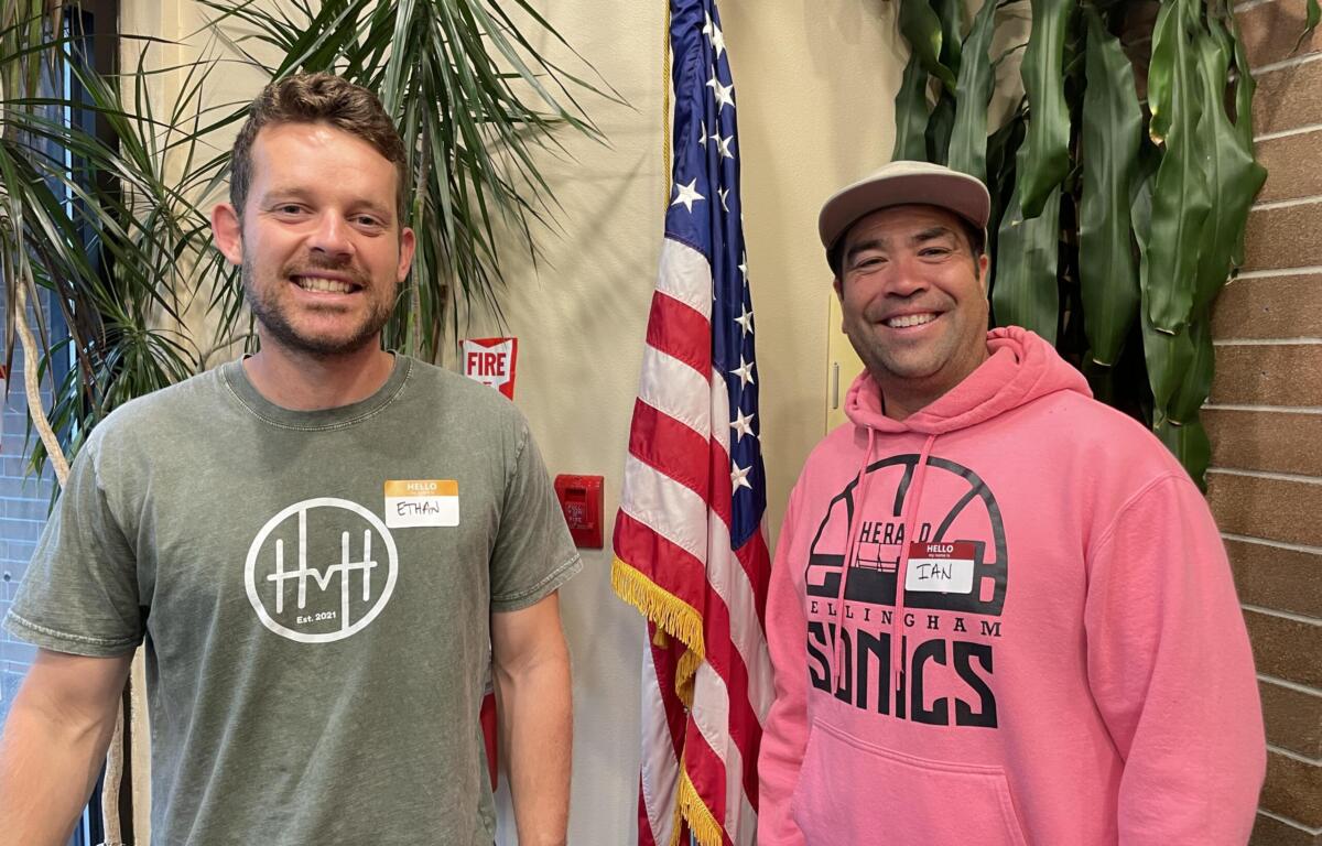 Two smiling men stand indoors with an American flag between them and greenery in the background, wearing name badges.