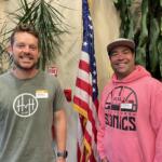 Two smiling men stand indoors with an American flag between them and greenery in the background, wearing name badges.