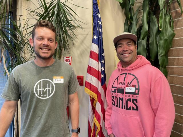Two smiling men stand indoors with an American flag between them and greenery in the background, wearing name badges.