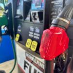 Red gasoline nozzle at a gas pump, with hoses and a payment terminal visible in the background.