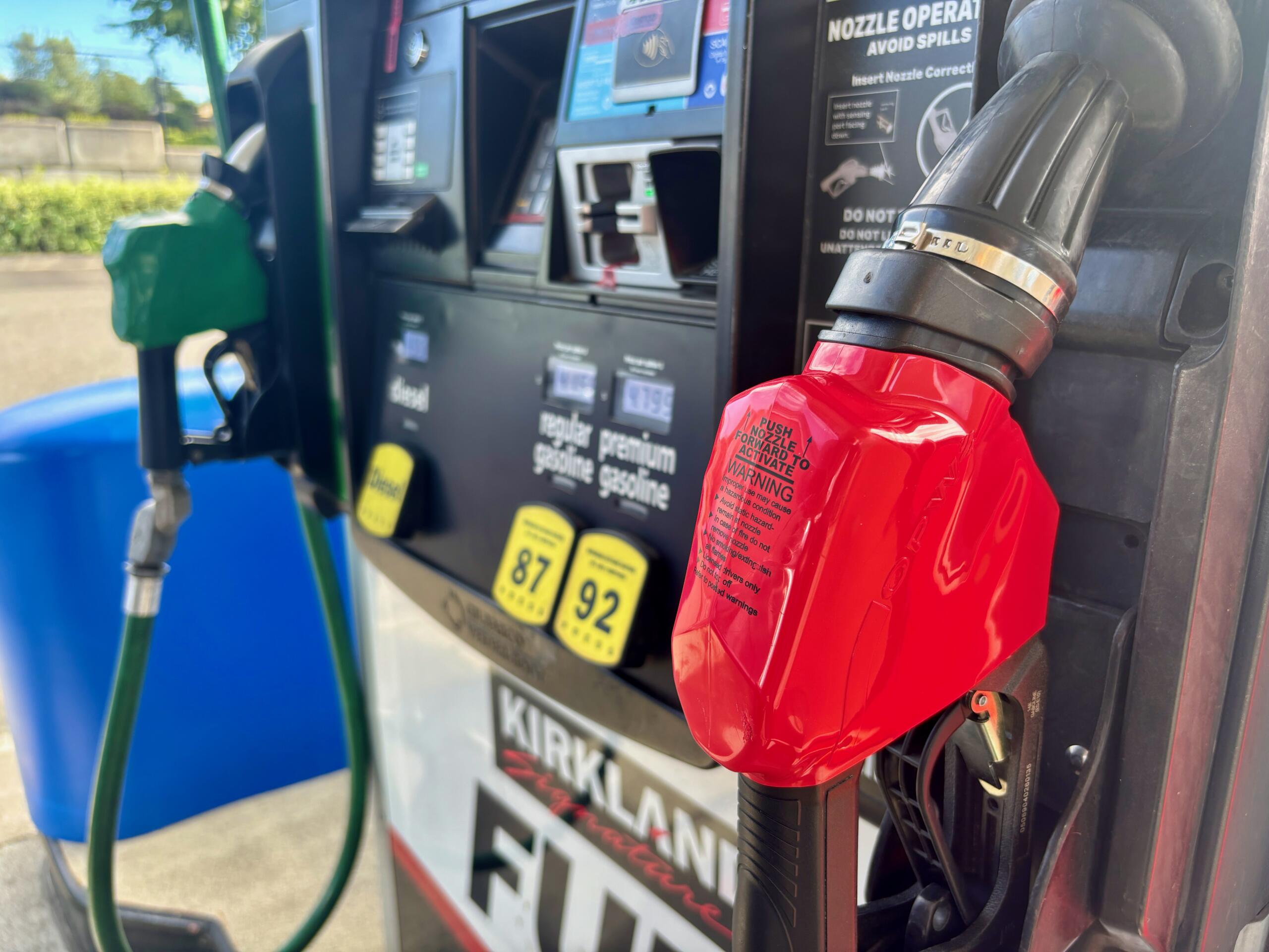 Red gasoline nozzle at a gas pump, with hoses and a payment terminal visible in the background.