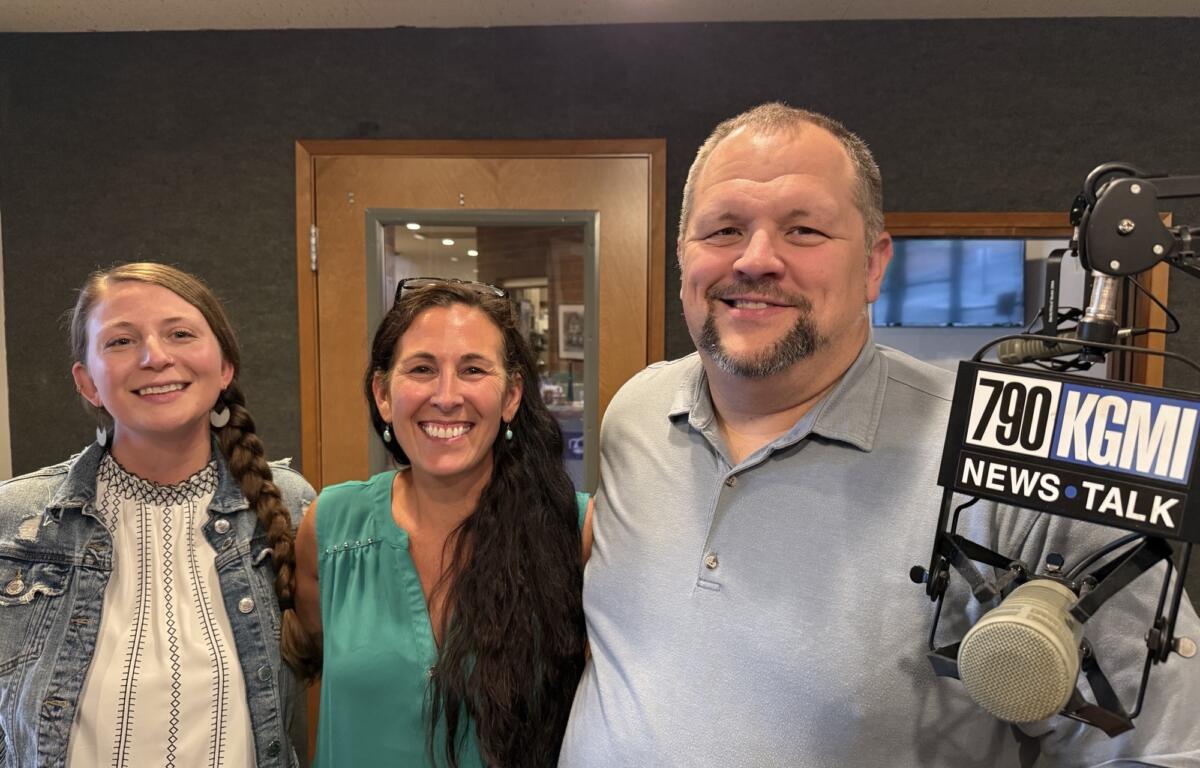 Three smiling colleagues posing in a radio studio beside a microphone, 790 KGM sign visible.