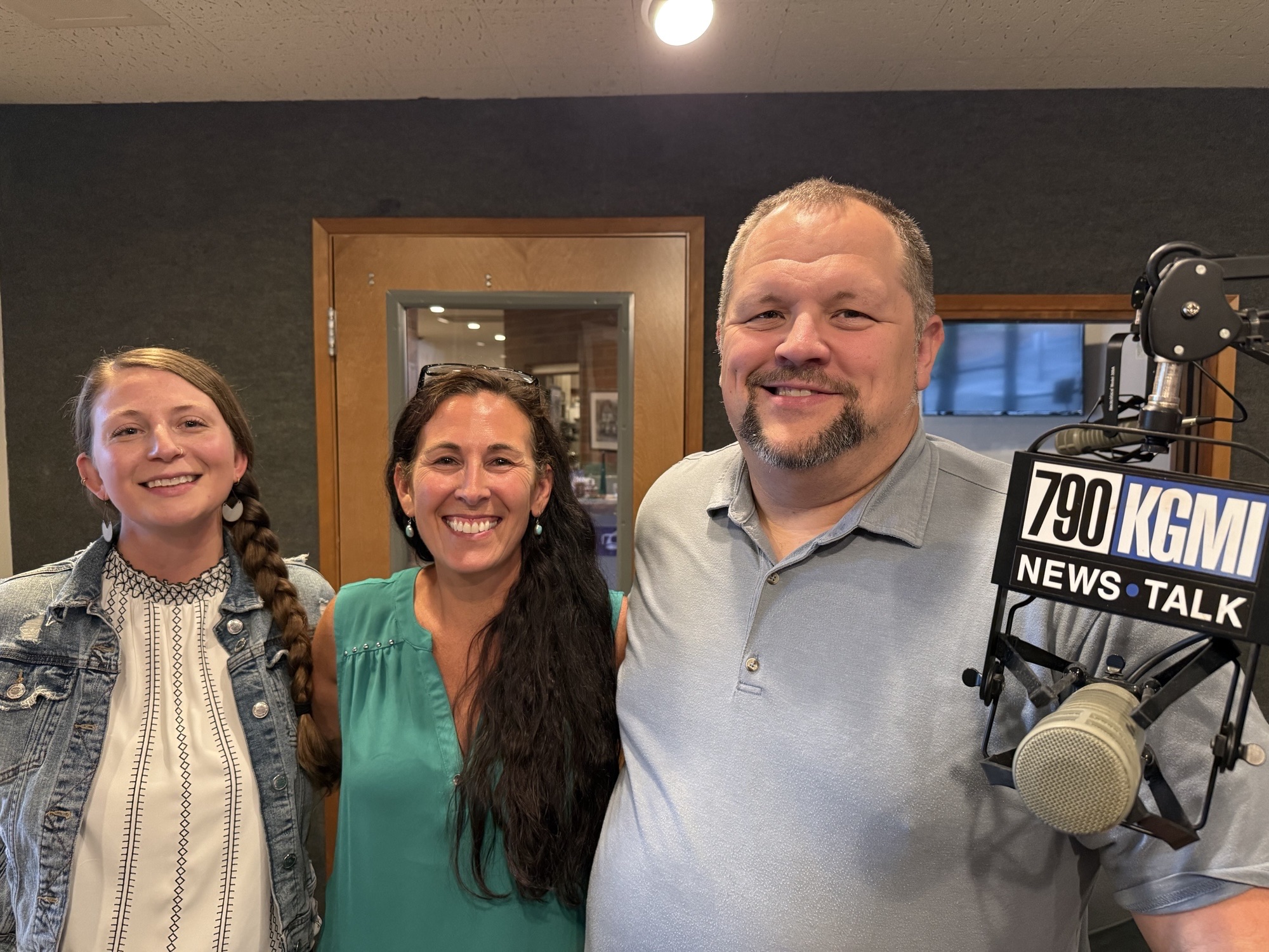 Three smiling colleagues posing in a radio studio beside a microphone, 790 KGM sign visible.