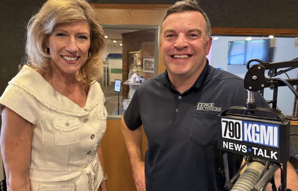 Smiling woman in a beige top and a man in a dark polo stand beside a radio microphone in a studio, with a 790 KGM News Talk sign nearby.