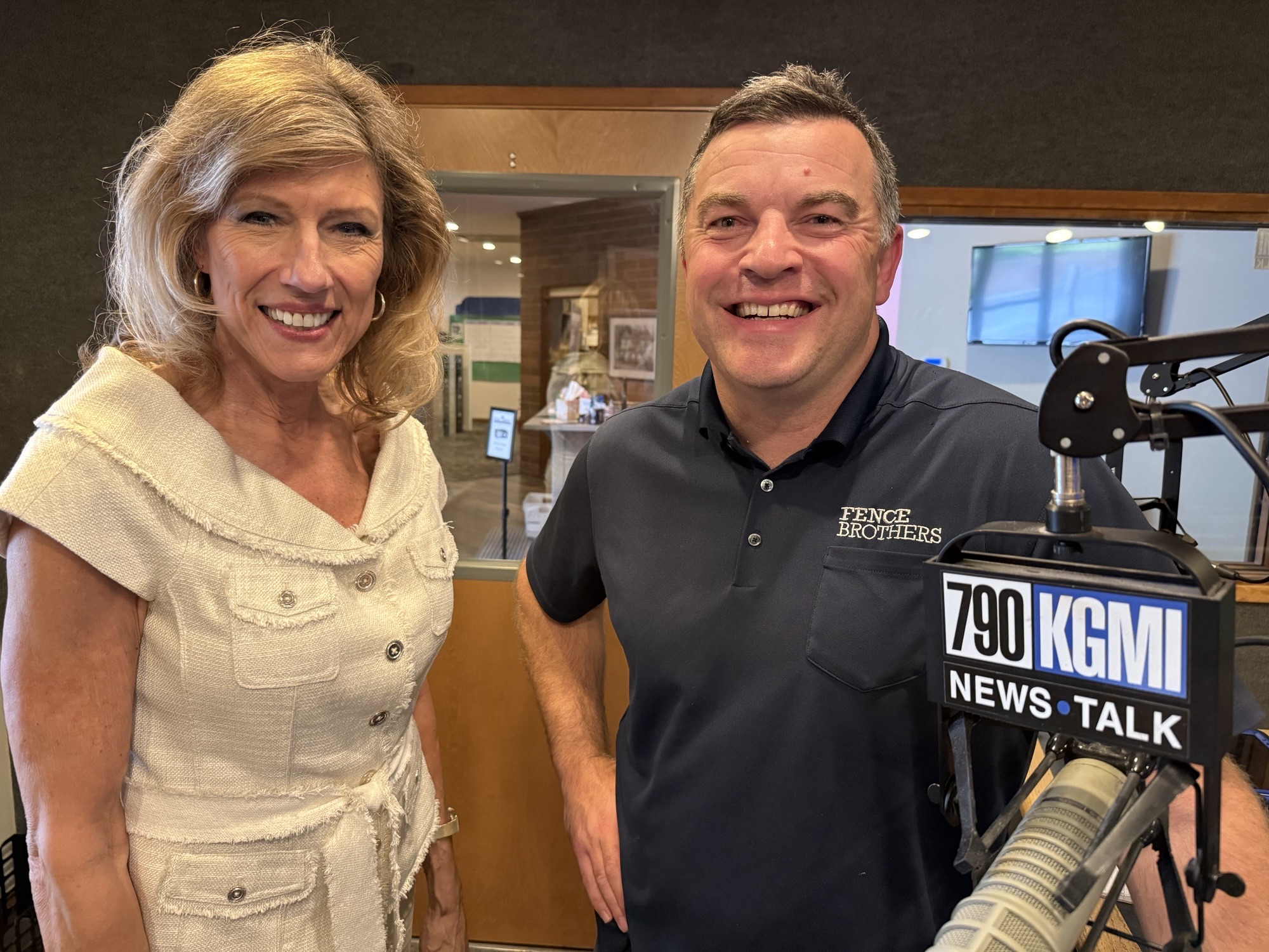 Smiling woman in a beige top and a man in a dark polo stand beside a radio microphone in a studio, with a 790 KGM News Talk sign nearby.