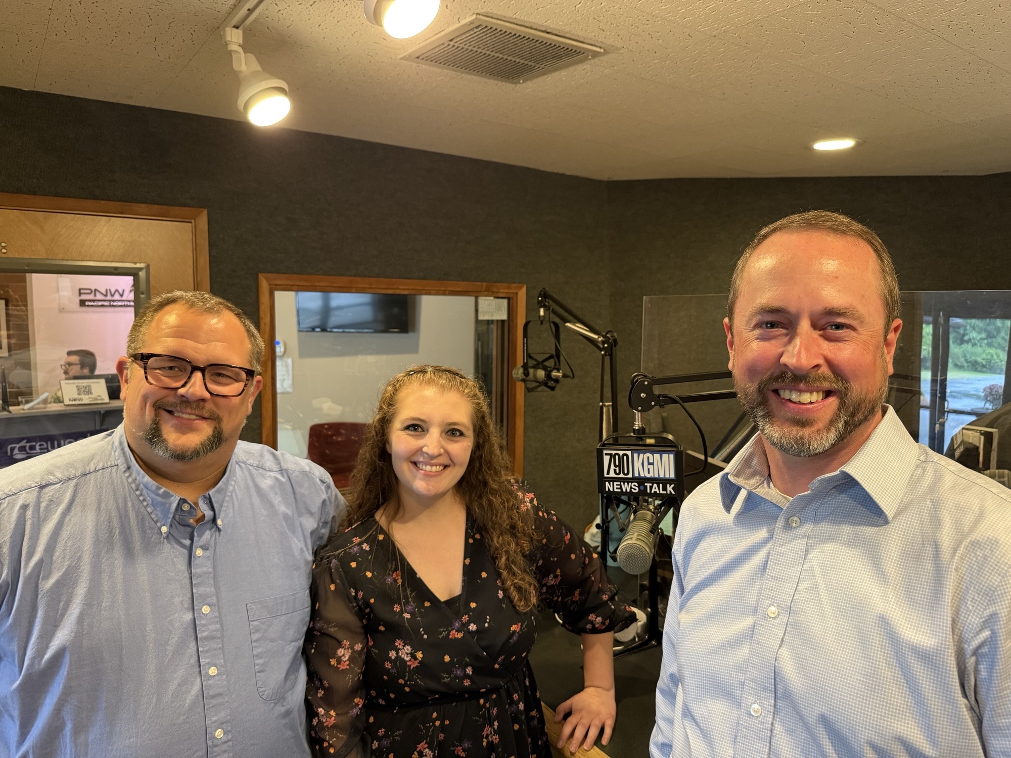 Three radio hosts pose for a selfie in a studio, smiling at the camera beside microphones and a '790 KGMI News Talk' sign on a mic stand.