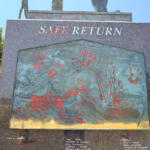 Monument plaque titled Safe Return, a bronze relief with red handprints and a sailing ship, outdoors by the water.