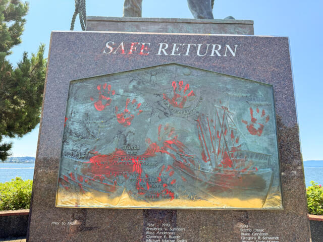 Monument plaque titled Safe Return, a bronze relief with red handprints and a sailing ship, outdoors by the water.