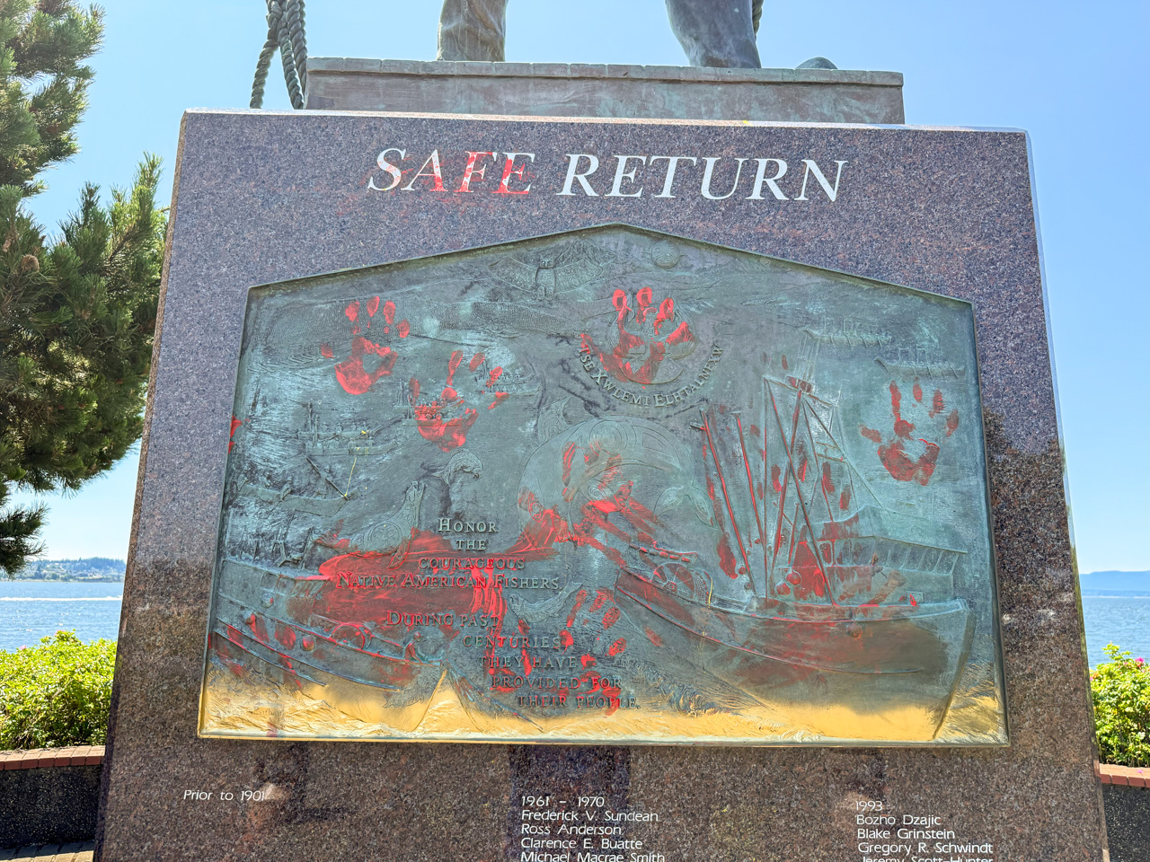 Monument plaque titled Safe Return, a bronze relief with red handprints and a sailing ship, outdoors by the water.