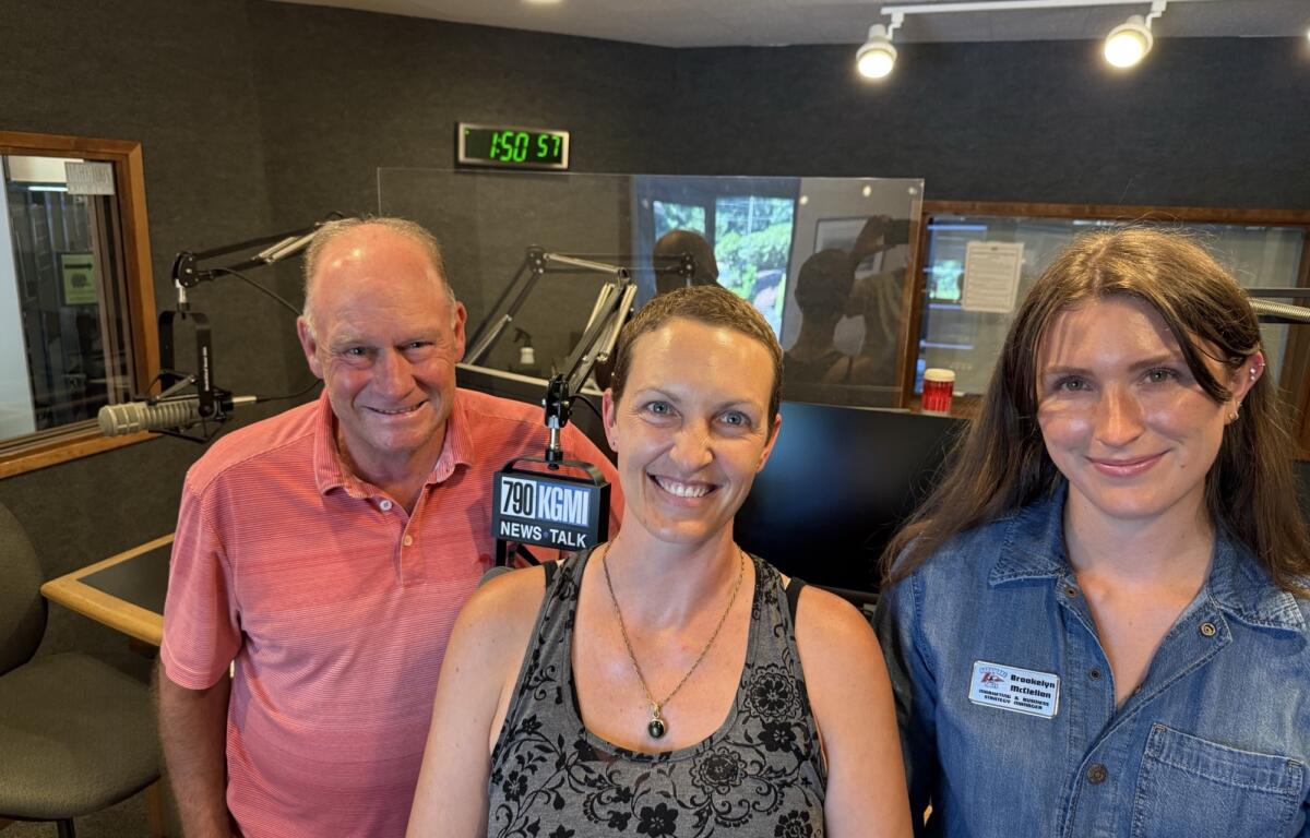 Three smiling people pose for a selfie in a radio studio with microphones and equipment around them, clock on wall behind.