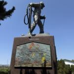 Bronze statue of a person holding a coiled rope atop a tall pedestal, set against a clear blue sky; plaque below features a textured relief with colorful markings and the inscription 'never to return'.