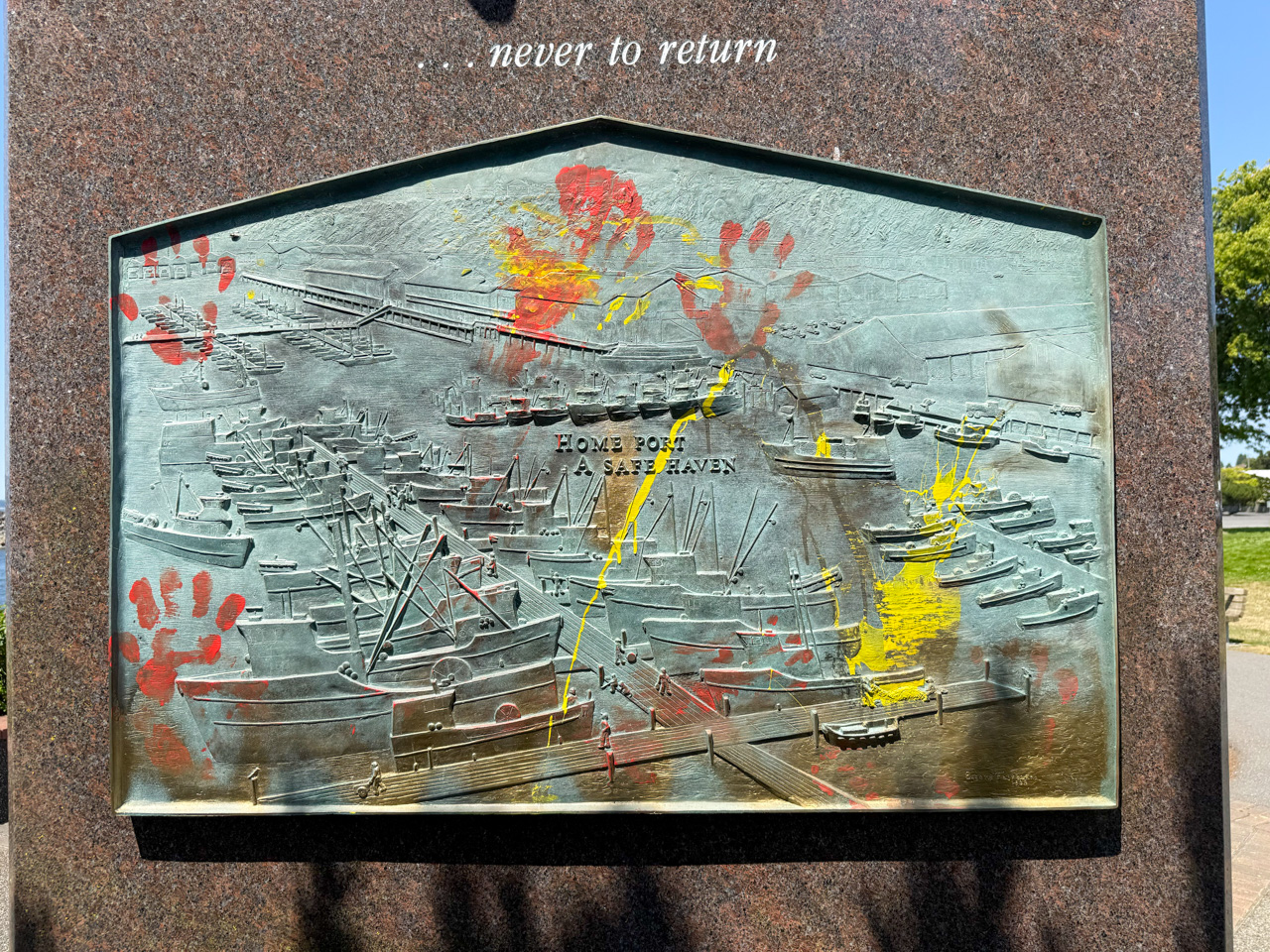 Relief plaque set in granite showing a crowded harbor with ships; reads 'HOME PORT A SAFE HAVEN' and has red/yellow handprints painted on it, plus the words '... never to return' above.