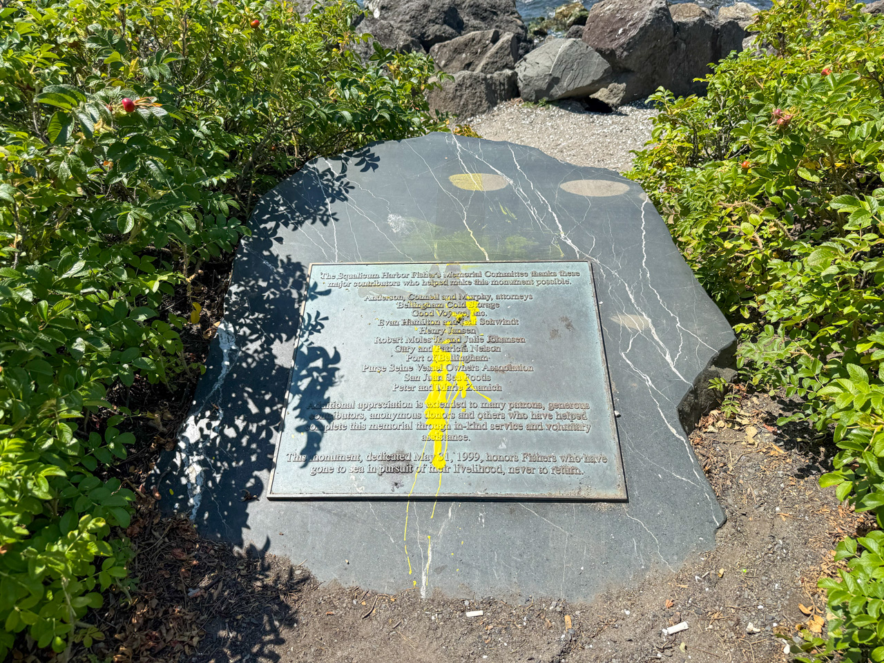 Bronze memorial plaque mounted on a dark slate rock at a rocky shoreline, flanked by green shrubs; yellow paint streaks cross the inscription, which commemorates fishermen who have gone to sea.