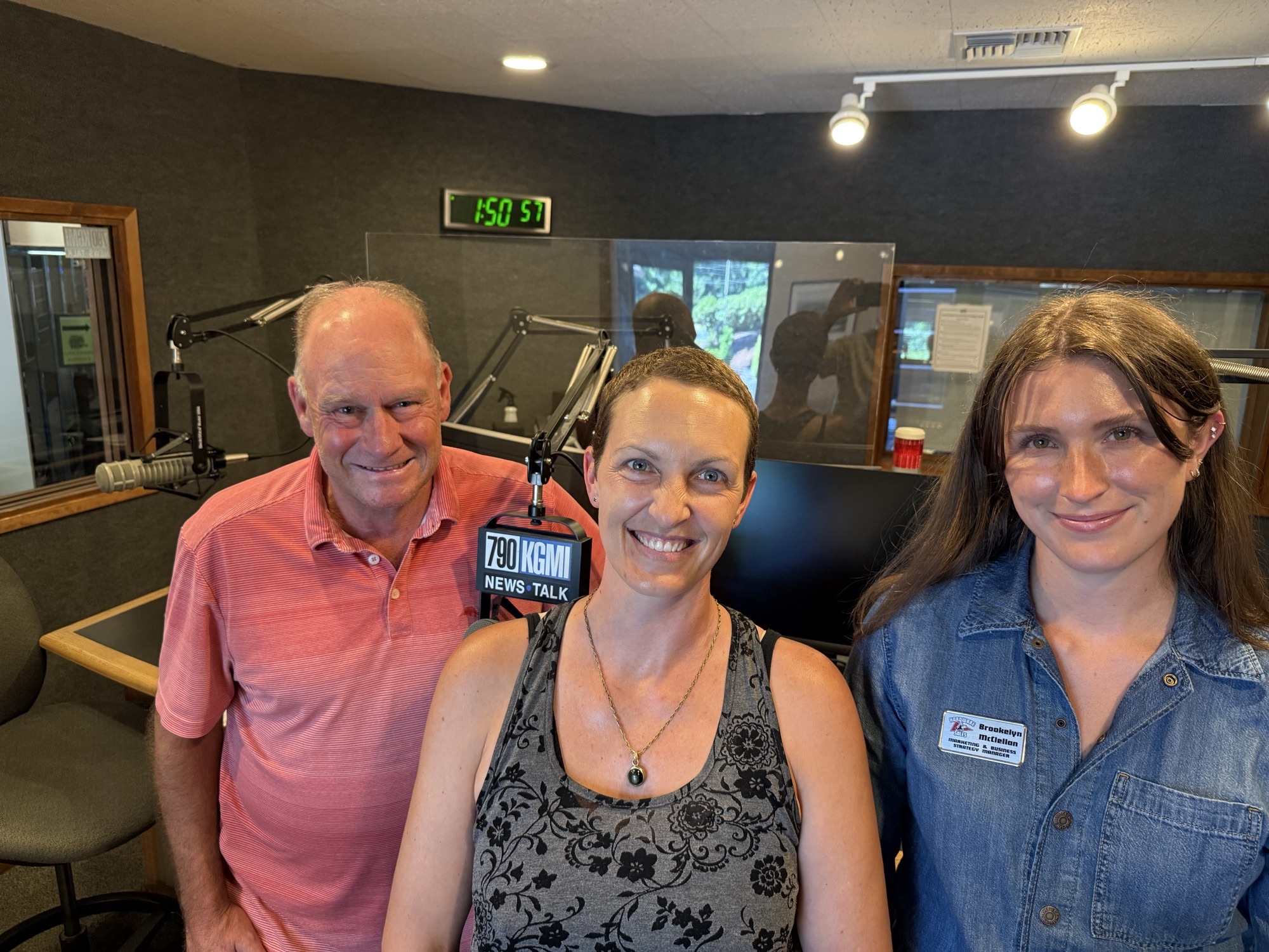 Three smiling people pose for a selfie in a radio studio with microphones and equipment around them, clock on wall behind.