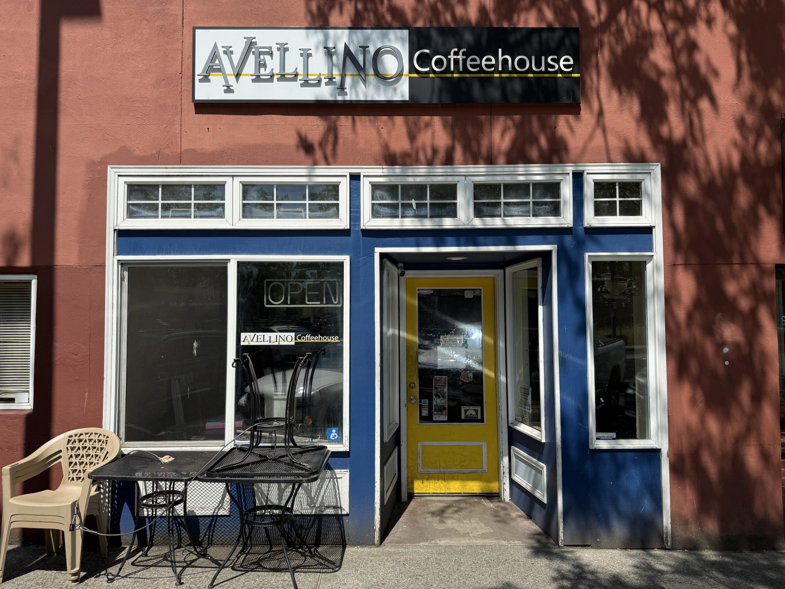 Front of Avellino Coffeehouse with blue storefront trim, yellow door, and outdoor metal tables and chairs under a tree-shadowed wall.