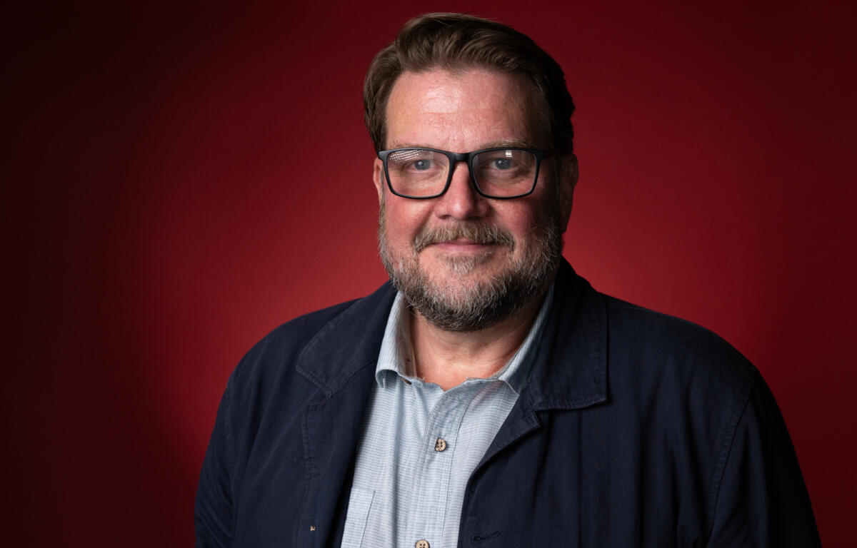 Friendly close-up portrait of a bearded man with glasses wearing a navy jacket against a red background, looking at the camera with a slight smile.