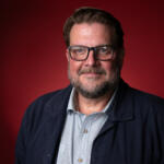 Friendly close-up portrait of a bearded man with glasses wearing a navy jacket against a red background, looking at the camera with a slight smile.