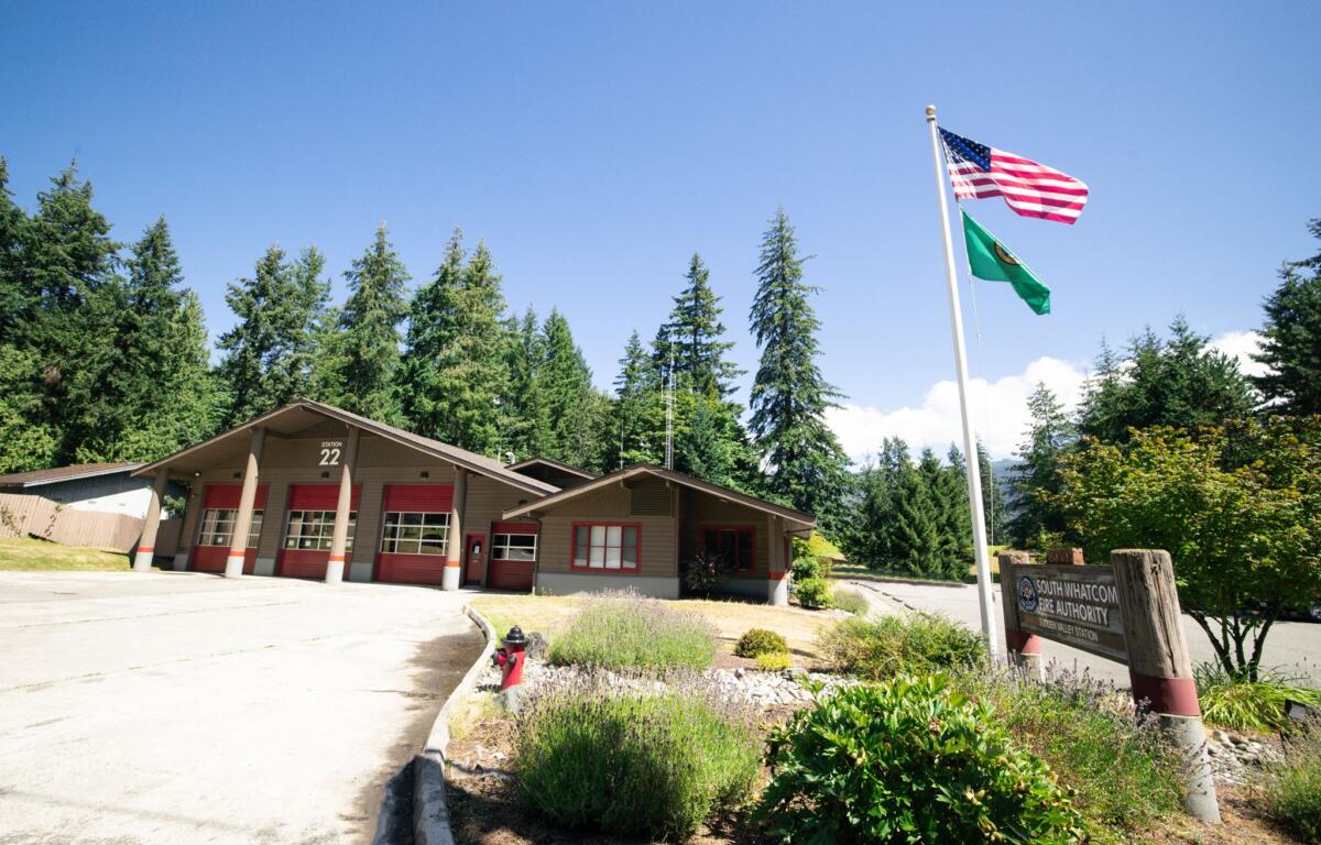 Fire station labeled Station 22 with red doors, beside a flagpole flying the U.S. flag and a green flag, surrounded by trees under a clear blue sky.