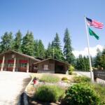 Fire station labeled Station 22 with red doors, beside a flagpole flying the U.S. flag and a green flag, surrounded by trees under a clear blue sky.