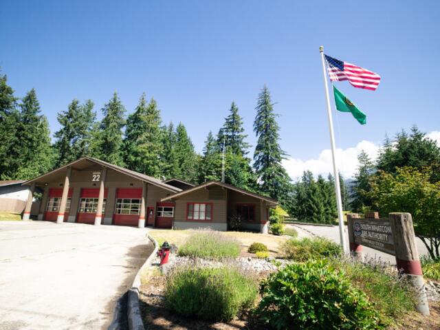 Fire station labeled Station 22 with red doors, beside a flagpole flying the U.S. flag and a green flag, surrounded by trees under a clear blue sky.