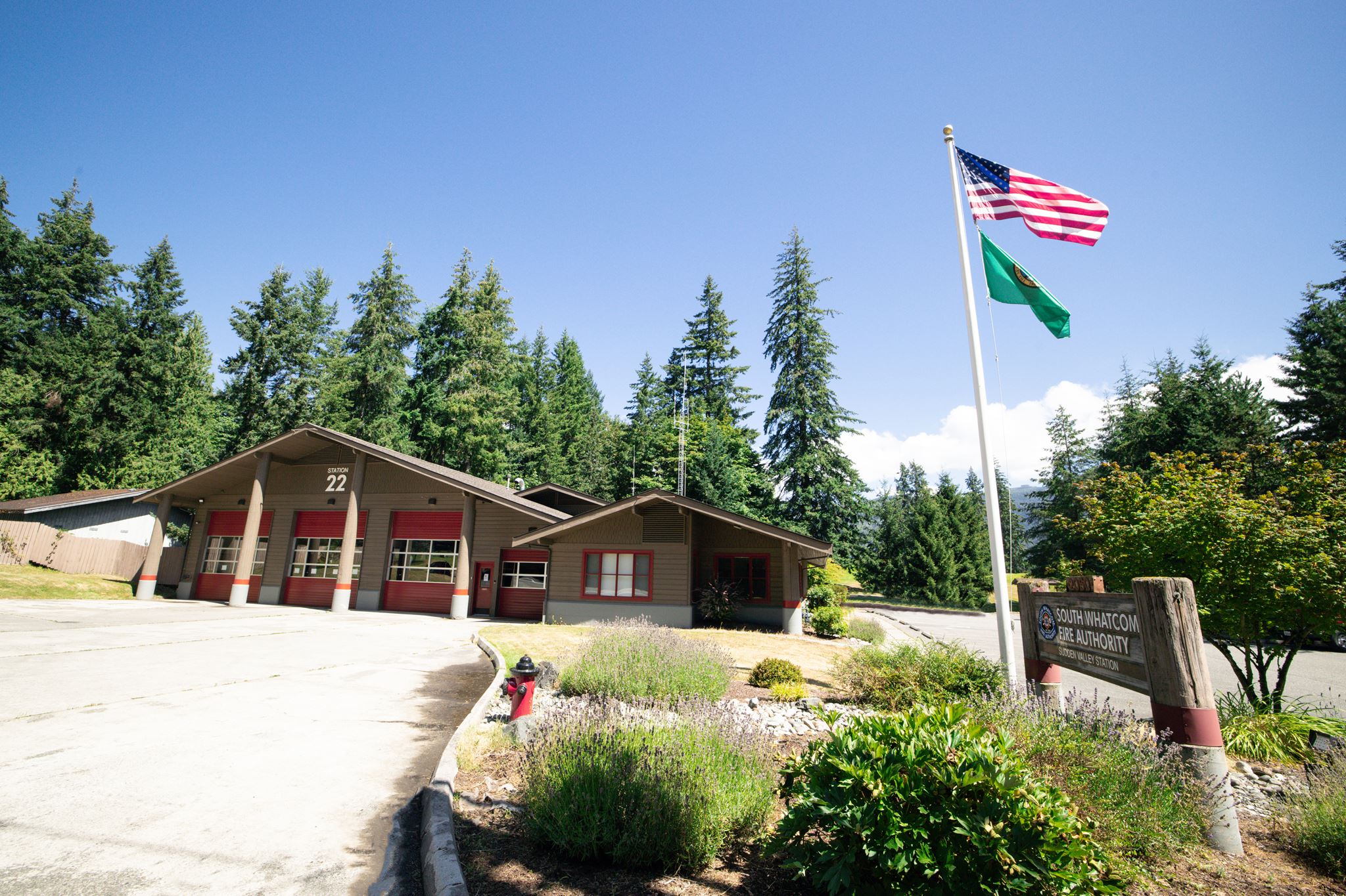 Fire station labeled Station 22 with red doors, beside a flagpole flying the U.S. flag and a green flag, surrounded by trees under a clear blue sky.