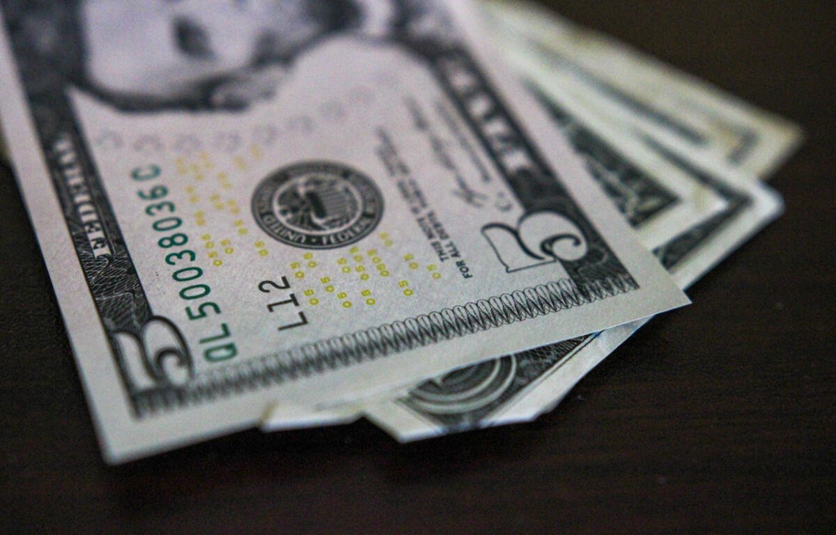 Close-up of several US five-dollar bills fanned on a dark surface with the Lincoln portrait area and serials visible on the front edge.