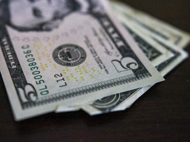 Close-up of several US five-dollar bills fanned on a dark surface with the Lincoln portrait area and serials visible on the front edge.