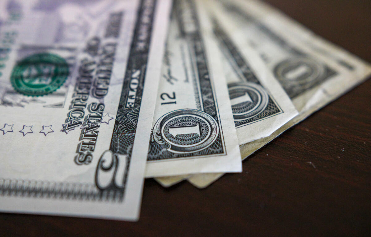 Fanned stack of US one-dollar bills on a dark wooden surface, showing the dollar sign and serials surrounding the front edges.