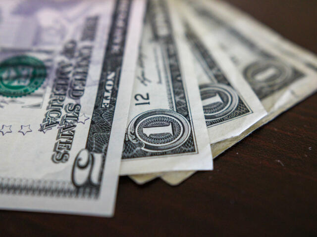 Fanned stack of US one-dollar bills on a dark wooden surface, showing the dollar sign and serials surrounding the front edges.