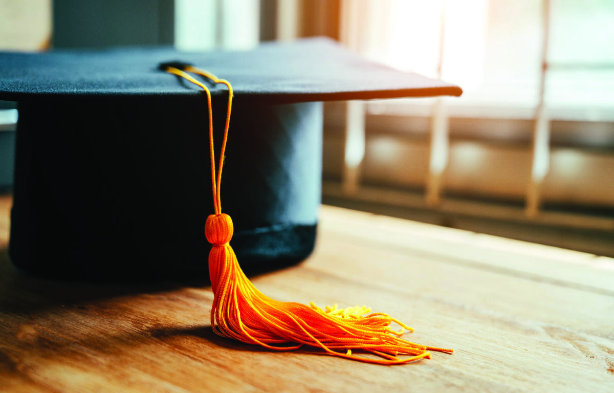 Close-up of a black graduation cap with an orange tassel resting on a wooden floor.