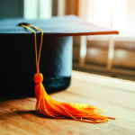 Close-up of a black graduation cap with an orange tassel resting on a wooden floor.