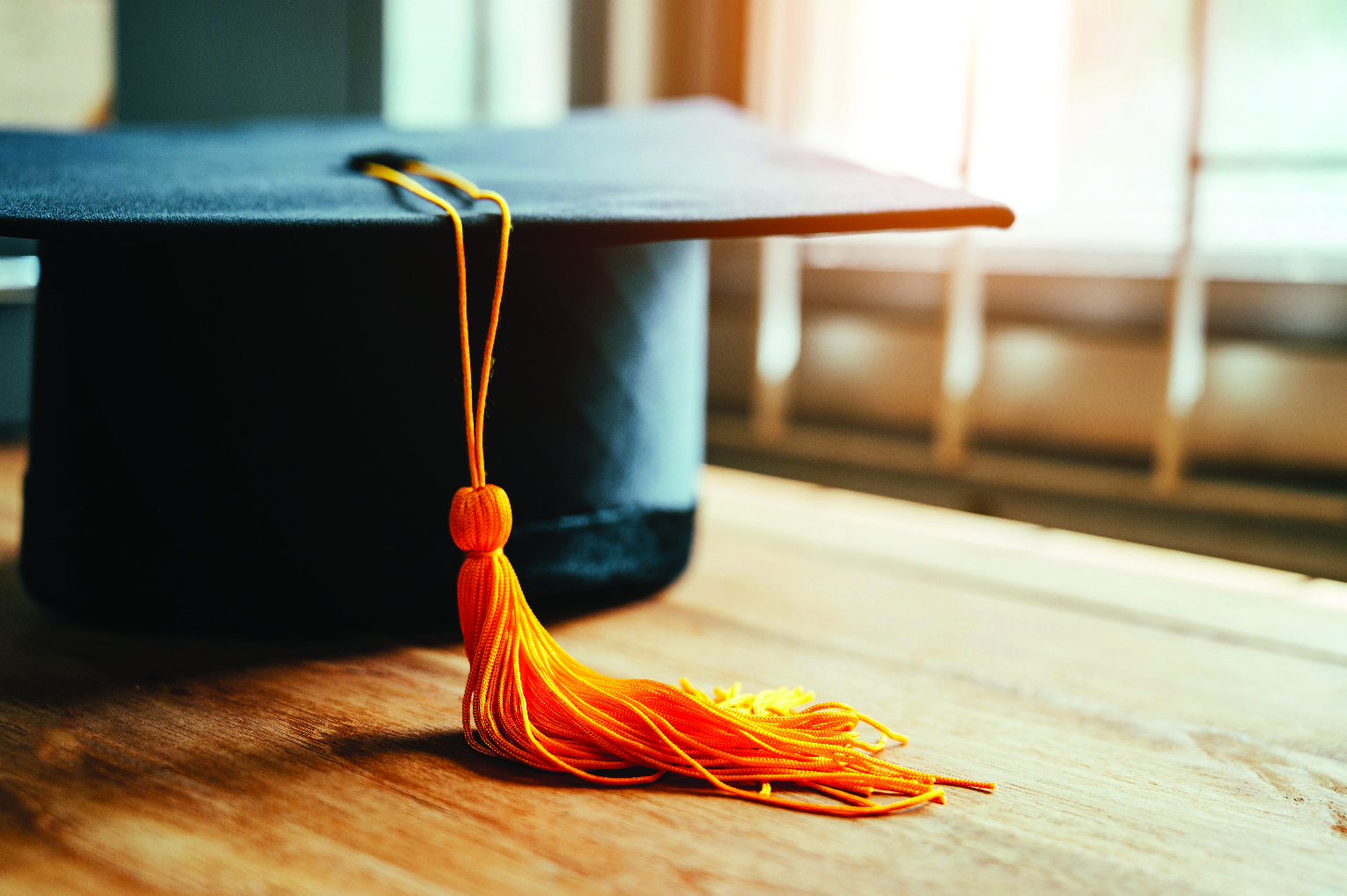 Close-up of a black graduation cap with an orange tassel resting on a wooden floor.