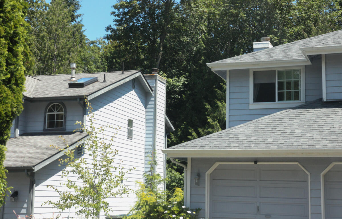 Two light-gray houses with attached garages set among lush trees and a clear blue sky in a suburban neighborhood run by sloped roofs and windows.
