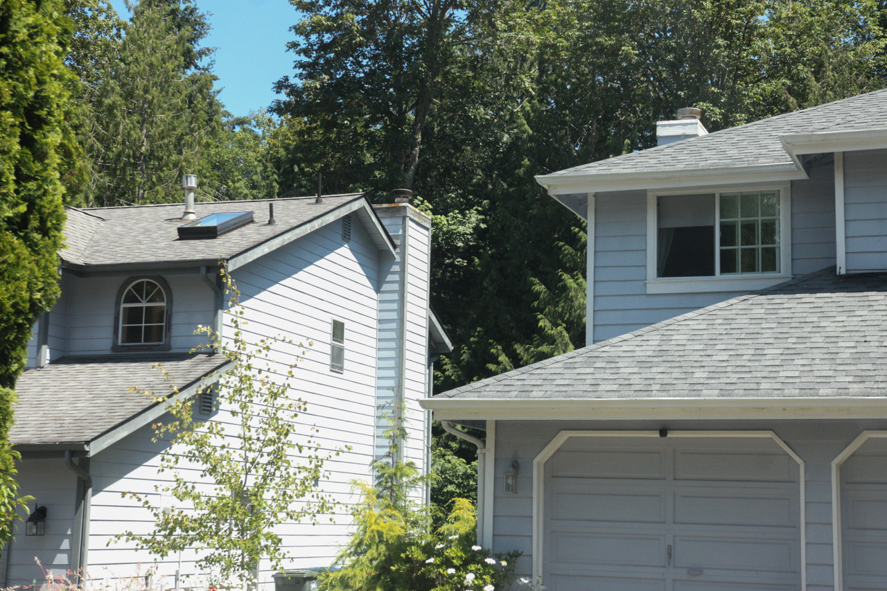 Two light-gray houses with attached garages set among lush trees and a clear blue sky in a suburban neighborhood run by sloped roofs and windows.