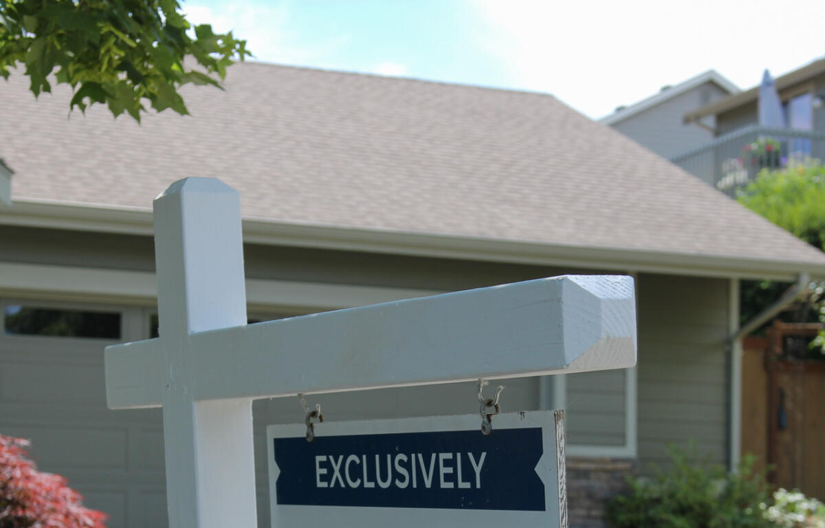 White wooden signpost with a blue 'EXCLUSIVELY' sign hanging in front of a suburban houseyard.