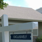White wooden signpost with a blue 'EXCLUSIVELY' sign hanging in front of a suburban houseyard.