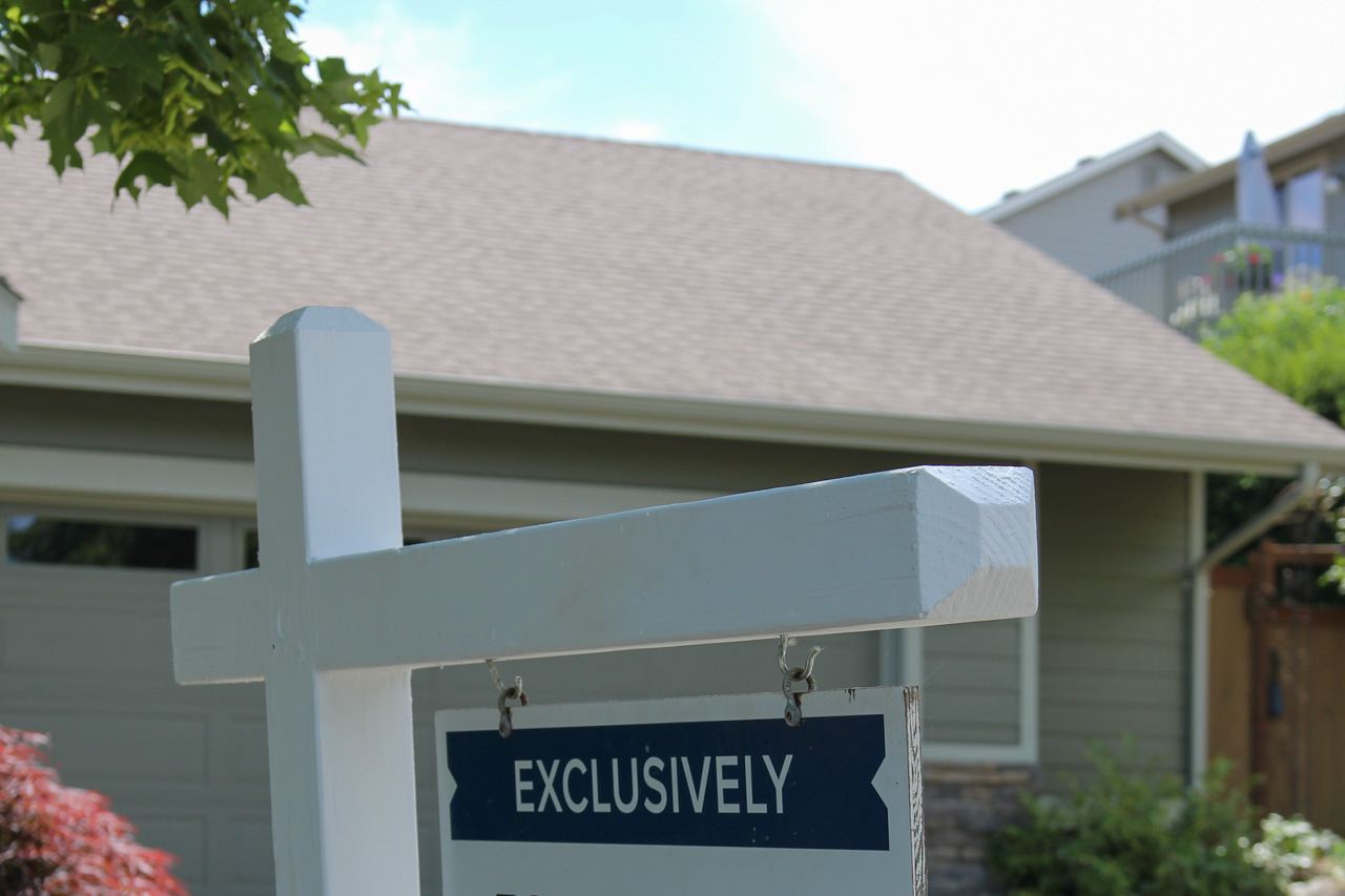 White wooden signpost with a blue 'EXCLUSIVELY' sign hanging in front of a suburban houseyard.