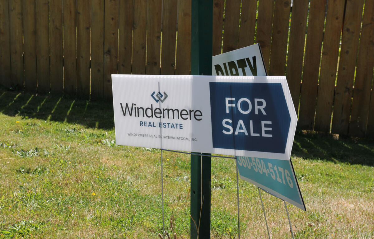 Windermere Real Estate 'For Sale' sign mounted on a post in a grassy yard, with a wooden fence in the background, visible logo and text.