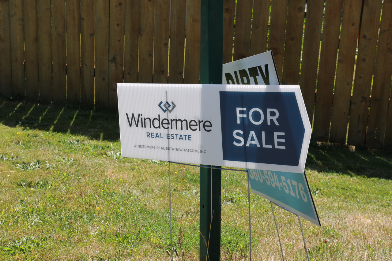 Windermere Real Estate 'For Sale' sign mounted on a post in a grassy yard, with a wooden fence in the background, visible logo and text.