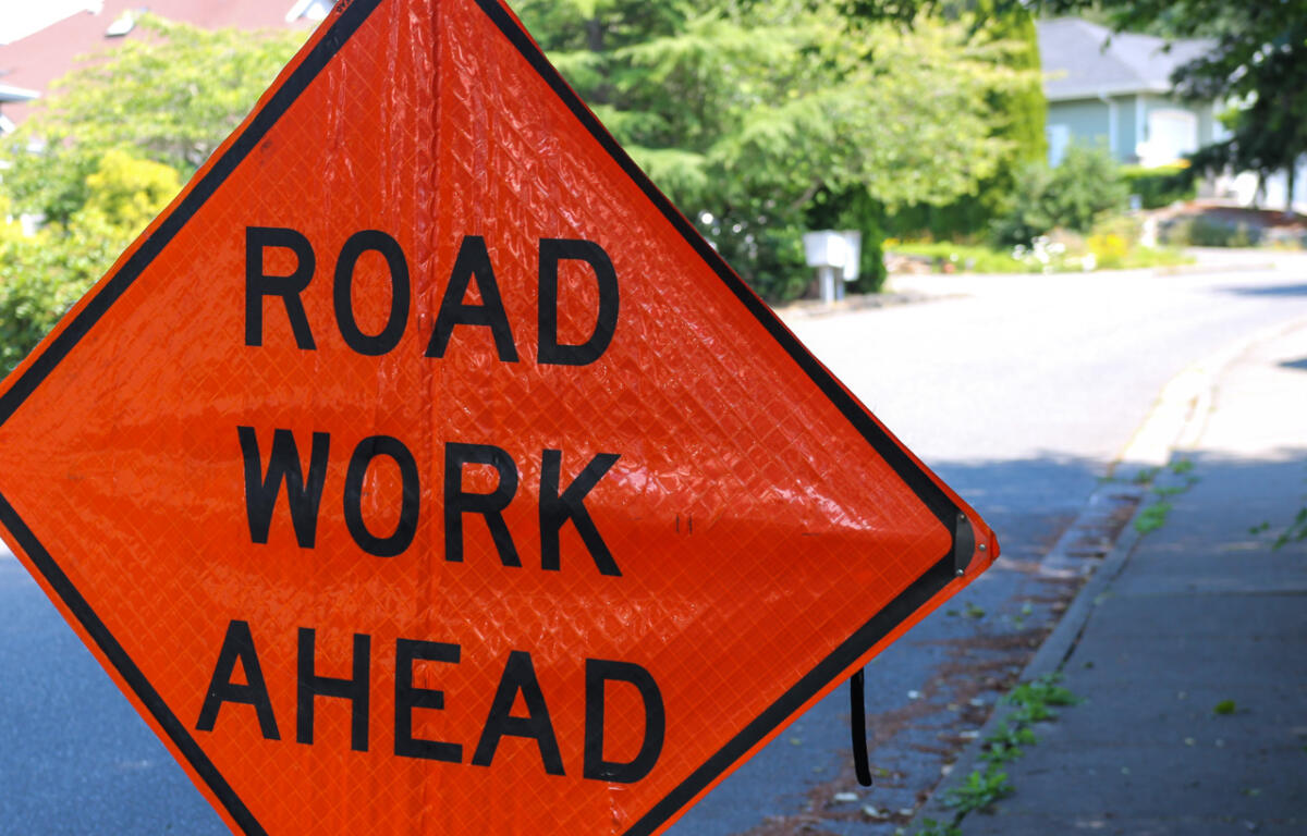 Orange diamond sign reading 'ROAD WORK AHEAD' on a residential street with greenery in the background.