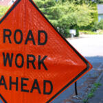 Orange diamond sign reading 'ROAD WORK AHEAD' on a residential street with greenery in the background.