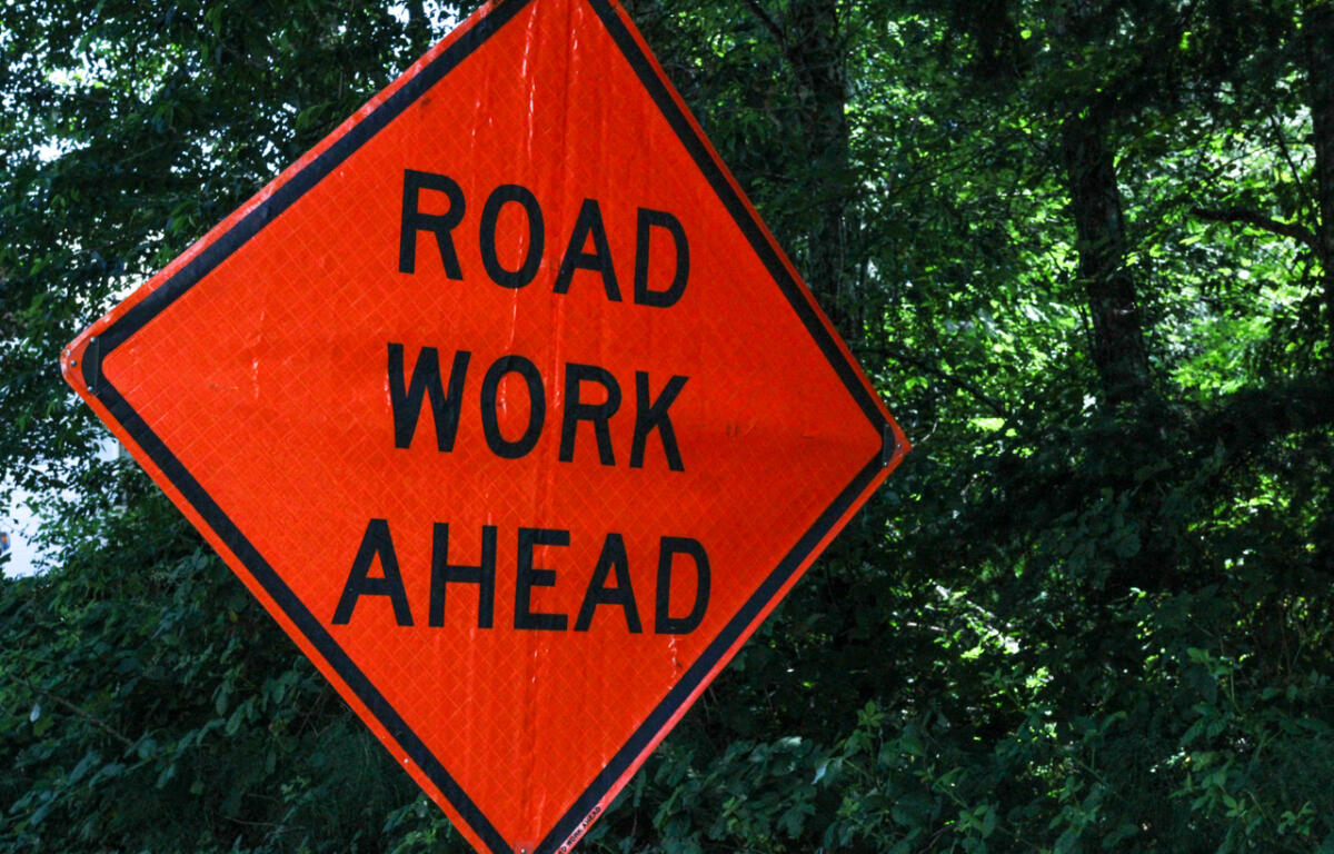 Orange diamond road work ahead sign with black text against green trees and bushes in the background.",