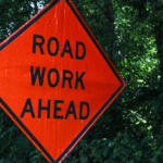 Orange diamond road work ahead sign with black text against green trees and bushes in the background.",